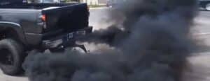 Black truck emitting dark smoke from exhaust on city street.